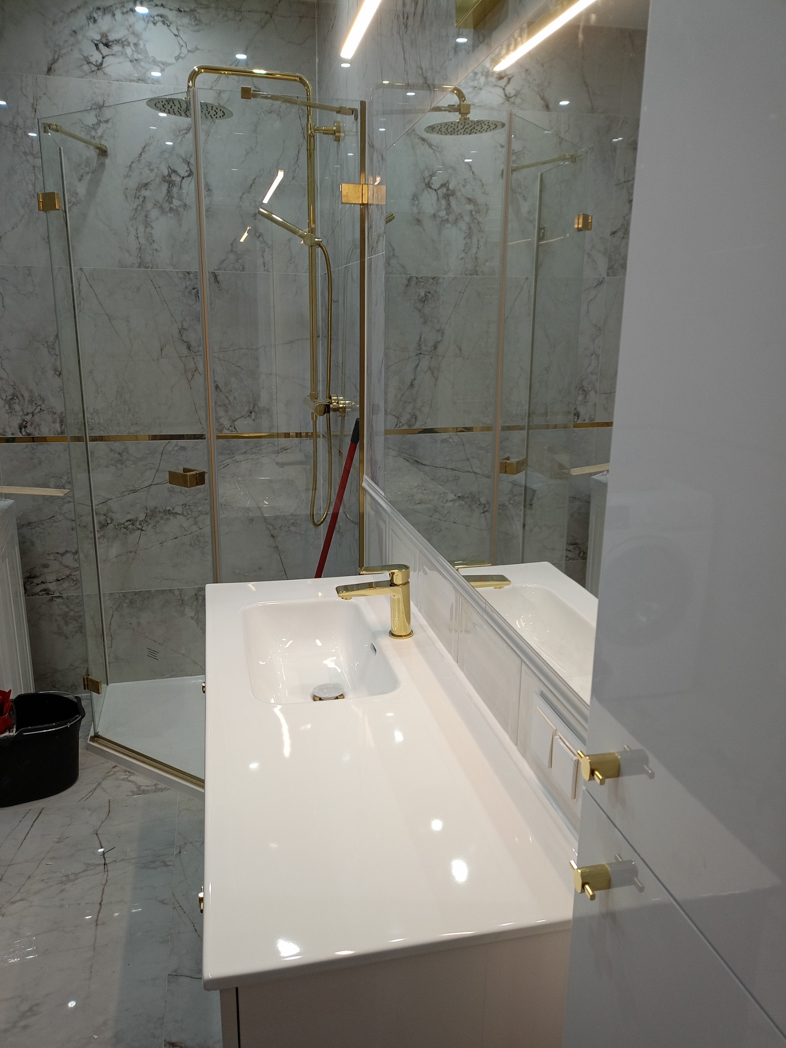 Nowoczesna łazienka z kabiną prysznicową typu walk-in z elementami w kolorze złotym, białą umywalką nablatową i marmurowymi płytkami na ścianach.