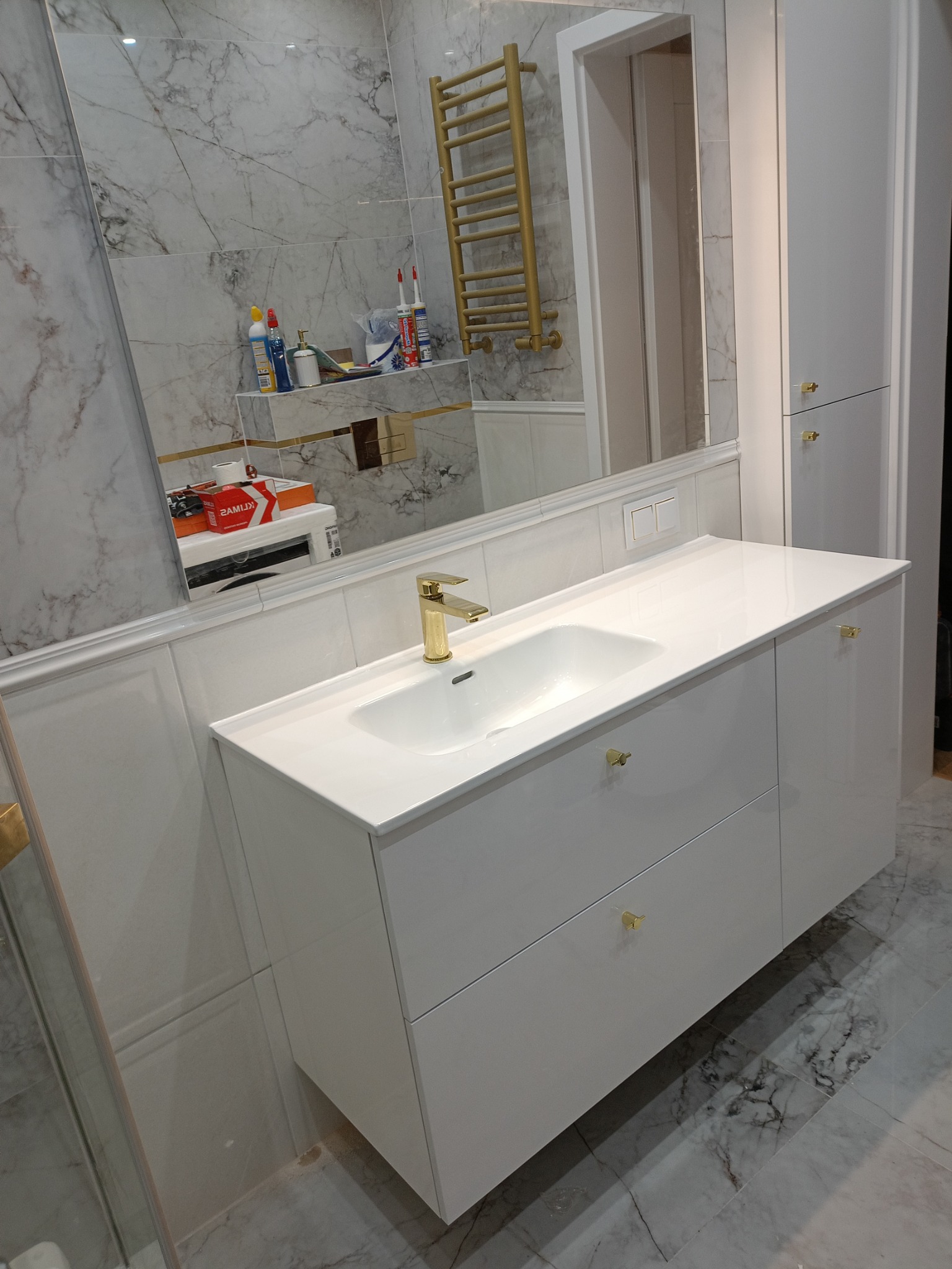 Nowoczesna łazienka z białą szafką pod umywalkę z złotymi uchwytami, marmurowe płytki na ścianach i podłodze, złoty grzejnik drabinkowy oraz duże lustro.