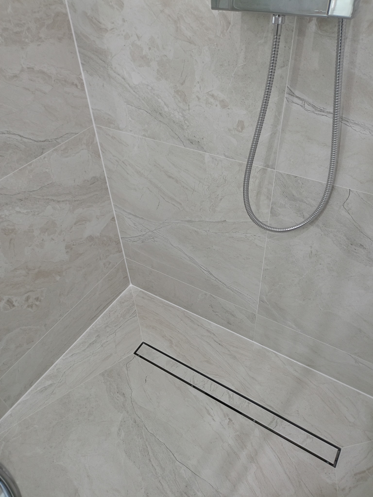 Szare marmurowe płytki podłogowe i ścienne w kabinie prysznicowej z odpływem liniowym i chromowaną słuchawką prysznicową.