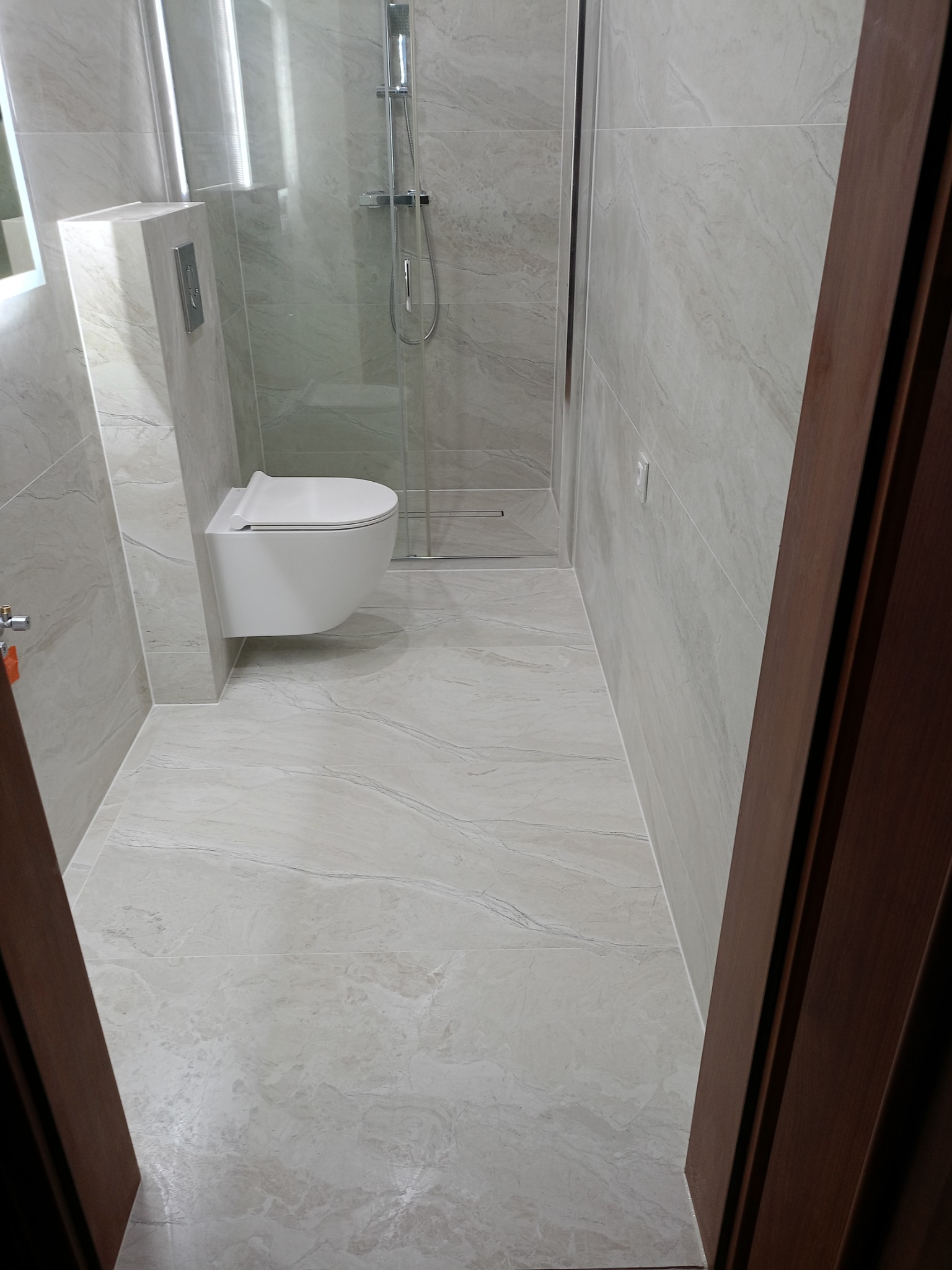 Nowoczesna łazienka z podwieszaną toaletą, kabiną prysznicową ze szklanymi drzwiami i płytkami imitującymi marmur na ścianach i podłodze, widok z perspektywy wejścia.