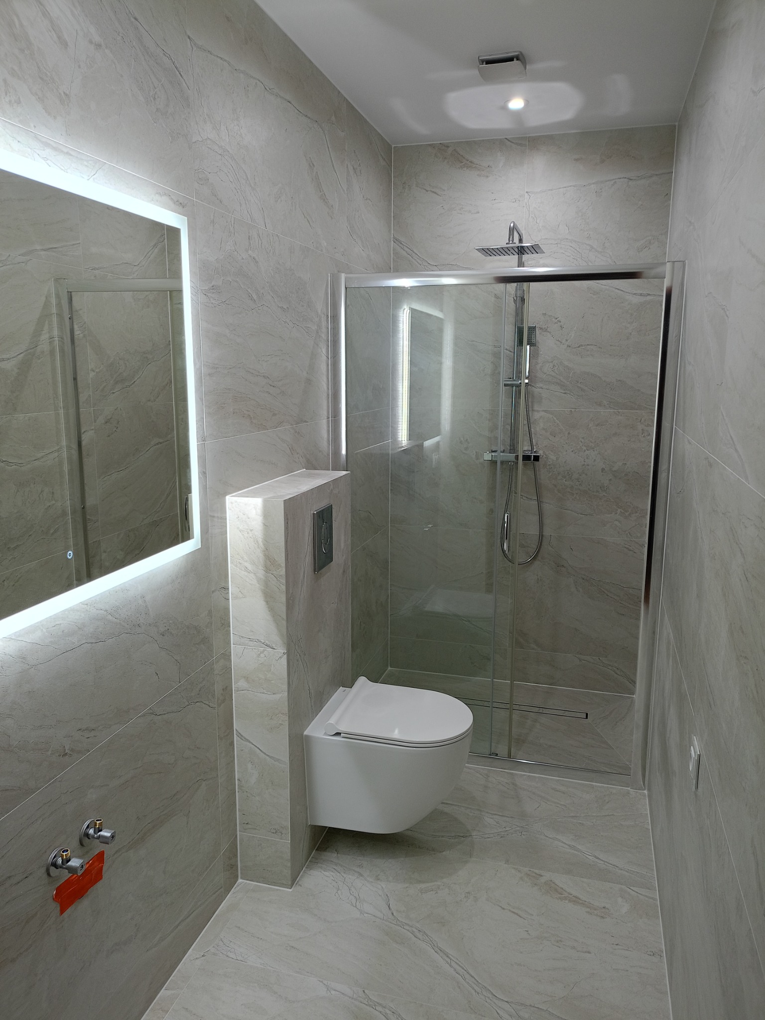 Nowoczesna łazienka z kabiną prysznicową typu walk-in, podwieszaną toaletą i lustrem z podświetleniem LED, wykończona płytkami imitującymi marmur.