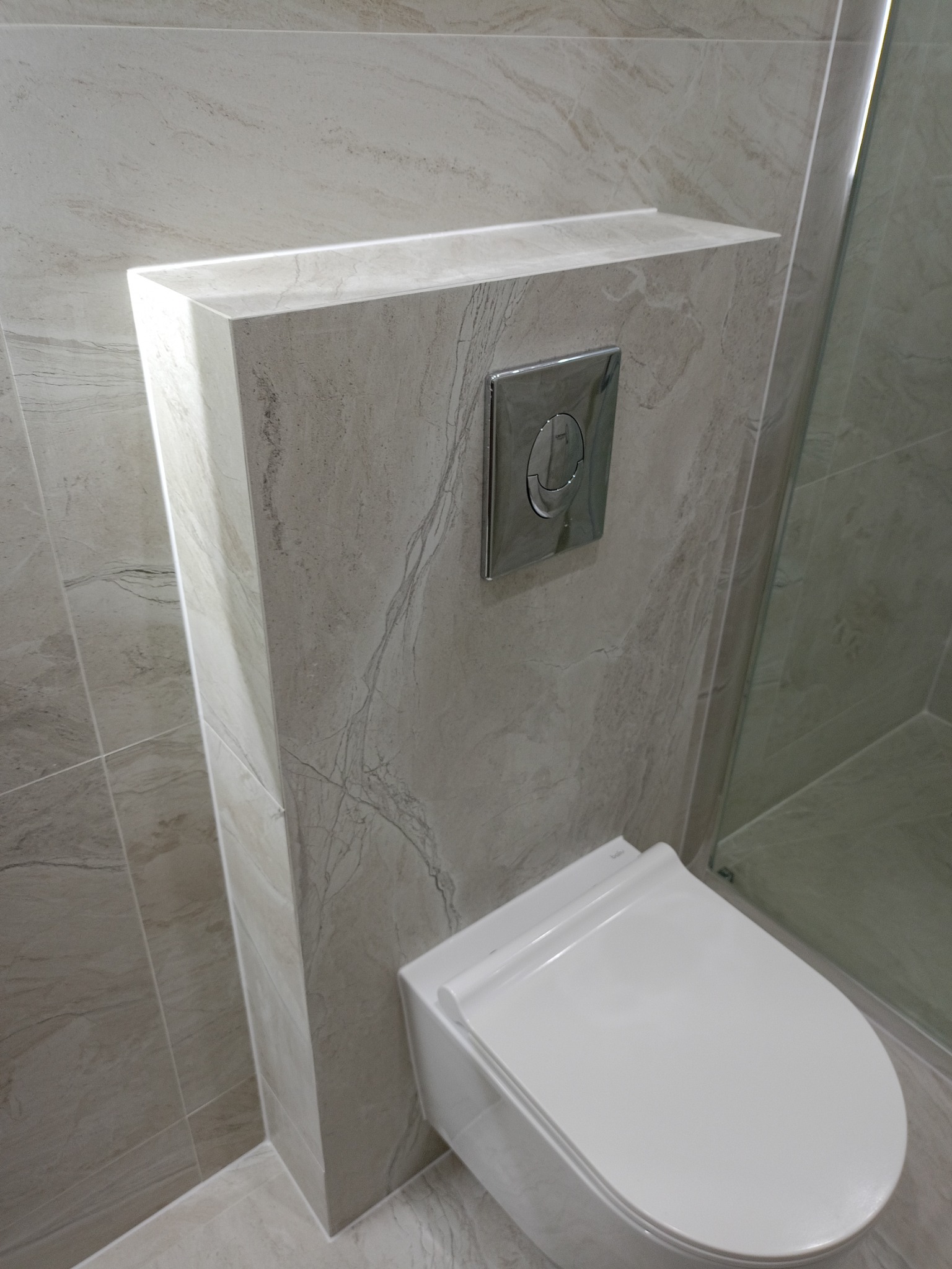 Podwieszana toaleta z zabudową geberit obłożona płytkami imitującymi marmur, chromowany przycisk spłukujący, kabina prysznicowa w tle.