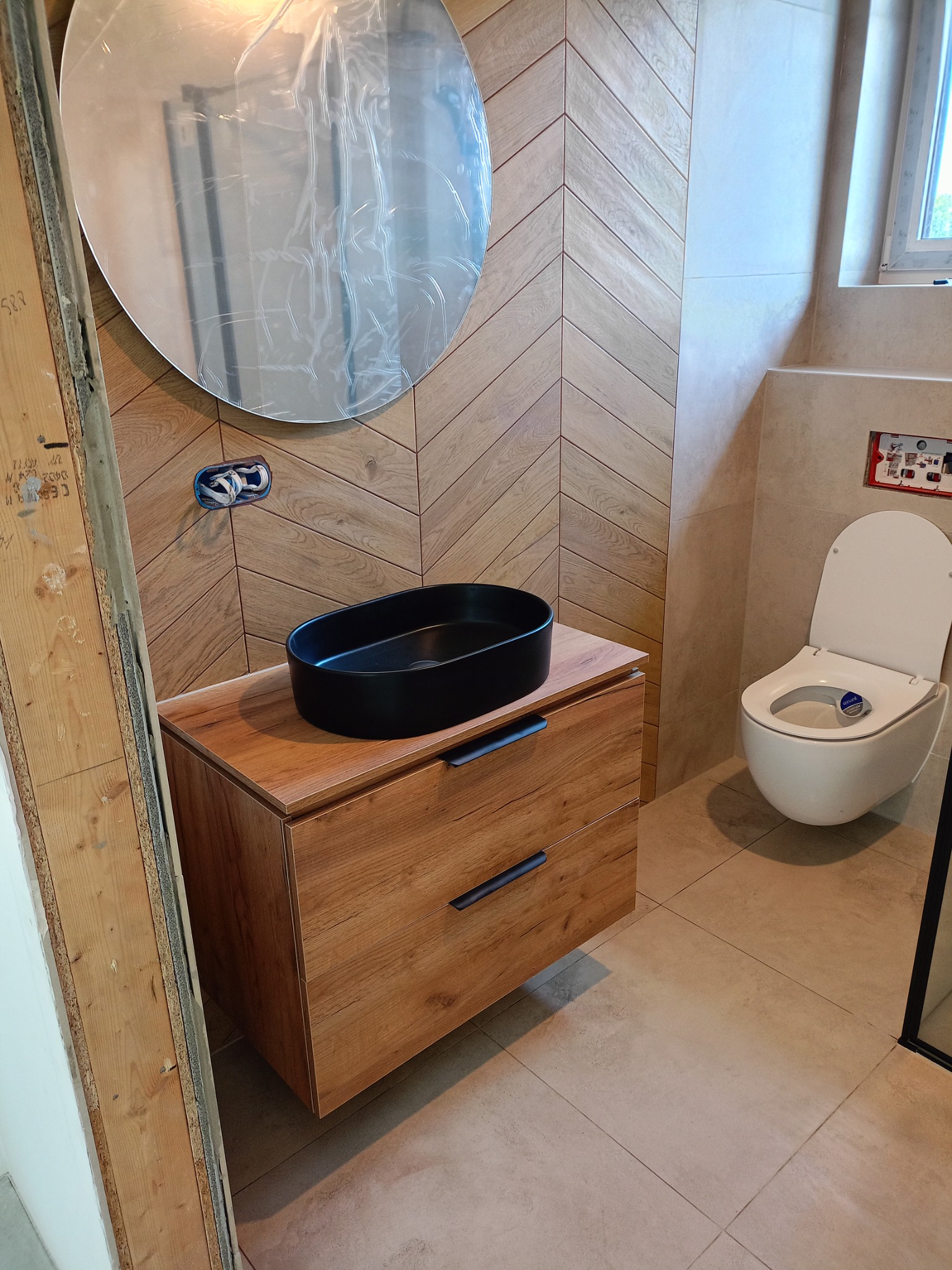 Nowoczesna łazienka z drewnianą szafką pod umywalką, owalną czarną umywalką nablatową, lustrem w folii ochronnej i toaletą podwieszaną. Na ścianie płytki w jodełkę.