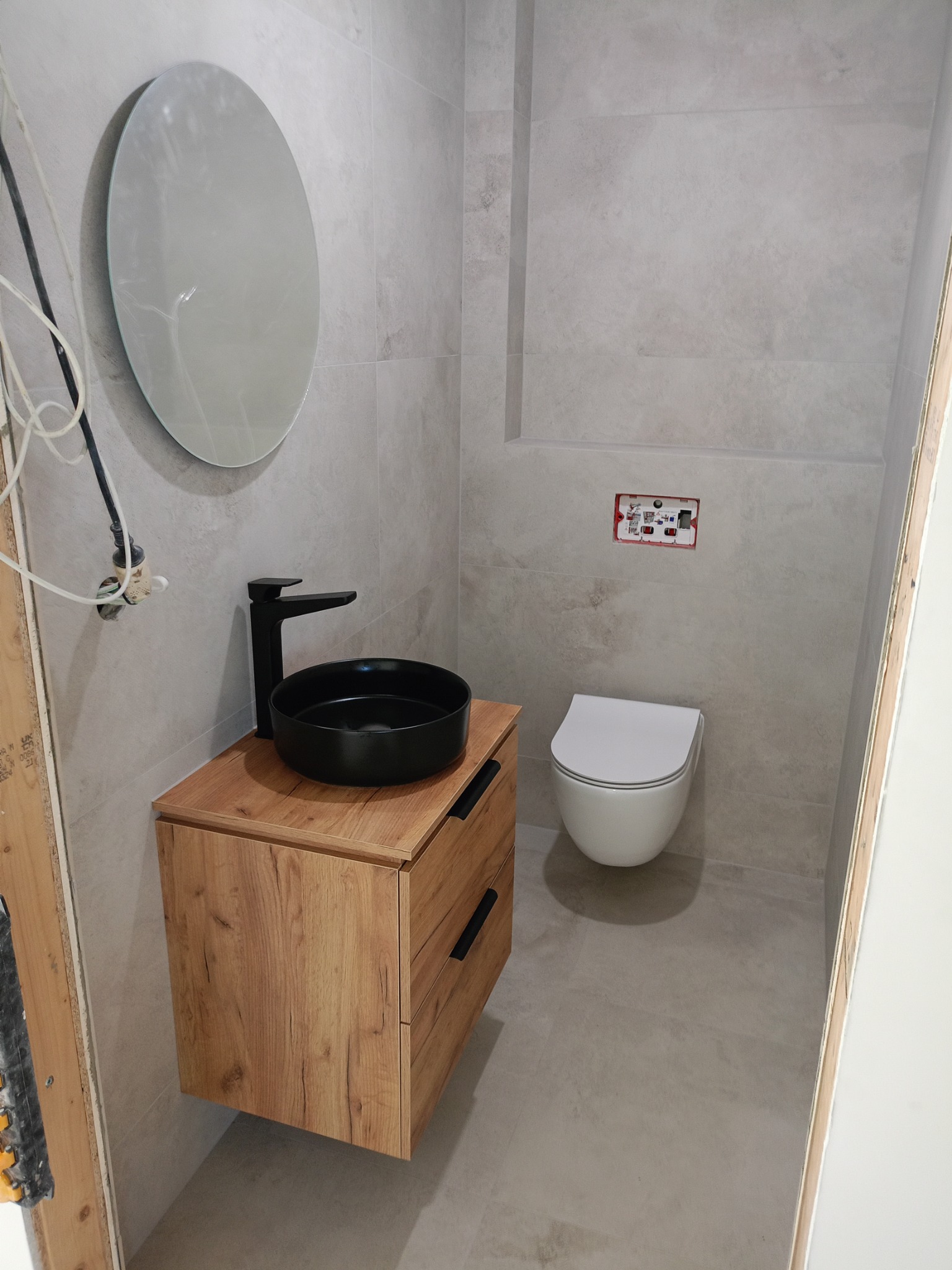 Nowoczesna łazienka w szarościach: okrągłe lustro, drewniana szafka z czarną umywalką i armaturą, podwieszana toaleta. Minimalistyczny design, świeżo po remoncie.