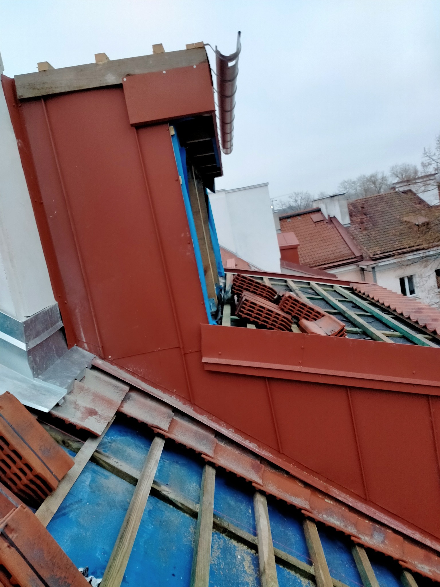Częściowo zdemontowany dach z widoczną konstrukcją drewnianą, zabezpieczoną niebieską folią, oraz stertą cegieł dachowych, z nową, czerwoną blachą trapezową na kominie.