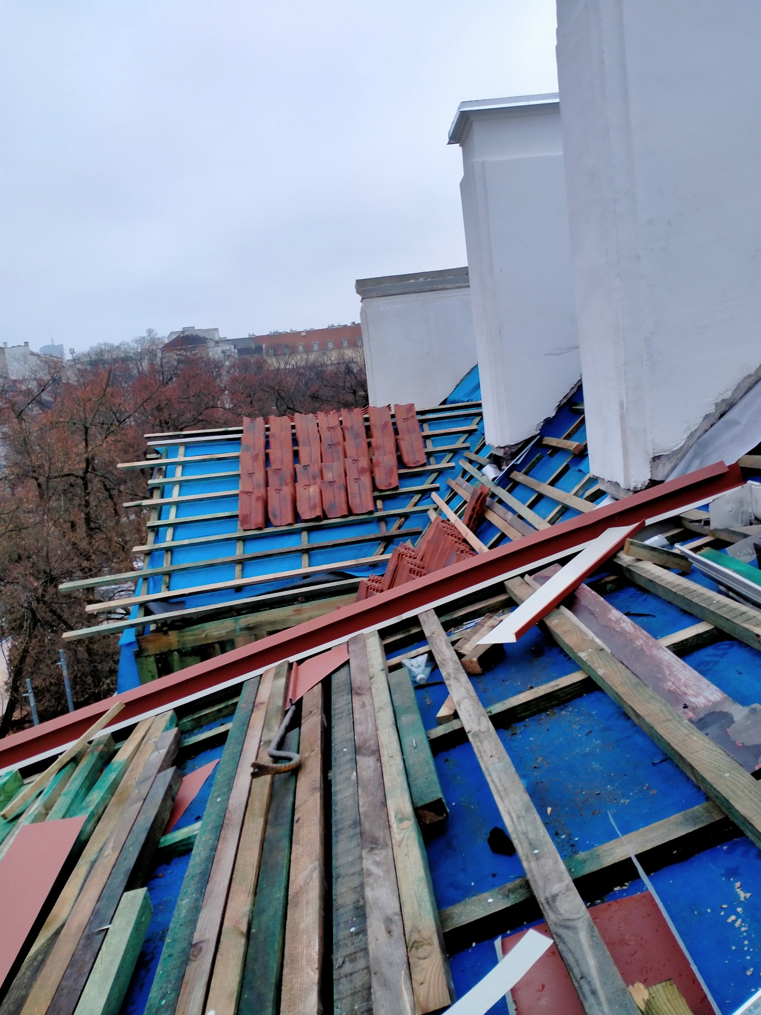 Widok z góry na konstrukcję dachu w trakcie budowy, z widocznymi łatami, kontrłatami, membraną w kolorze niebieskim oraz ułożonymi dachówkami w rzędach, a w tle budynki miejskie.