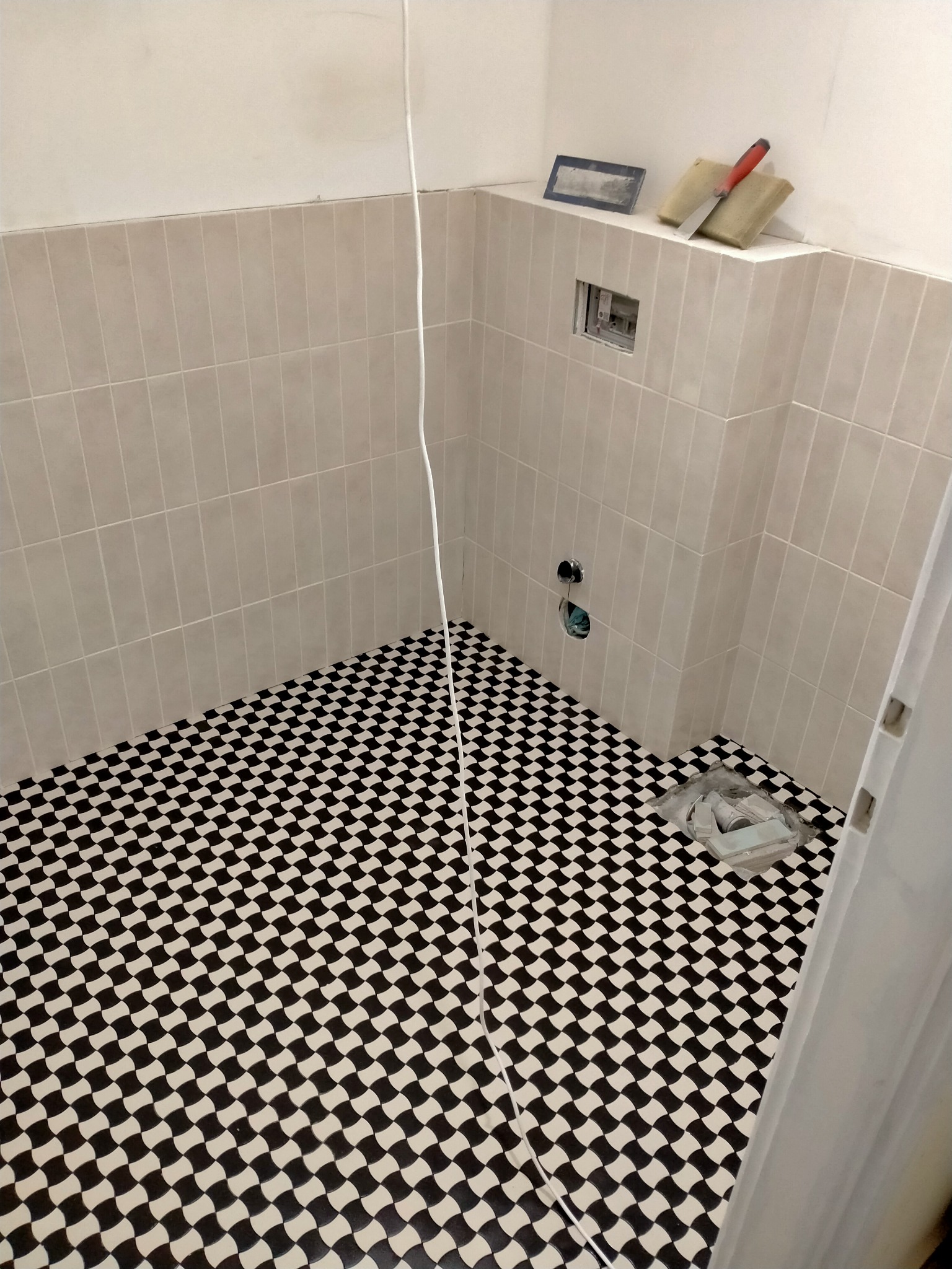 Wnętrze łazienki w trakcie remontu z ułożoną czarno-białą mozaiką podłogową, częściowo wykończone ściany z jasnymi płytkami, widoczne narzędzia i materiały budowlane.