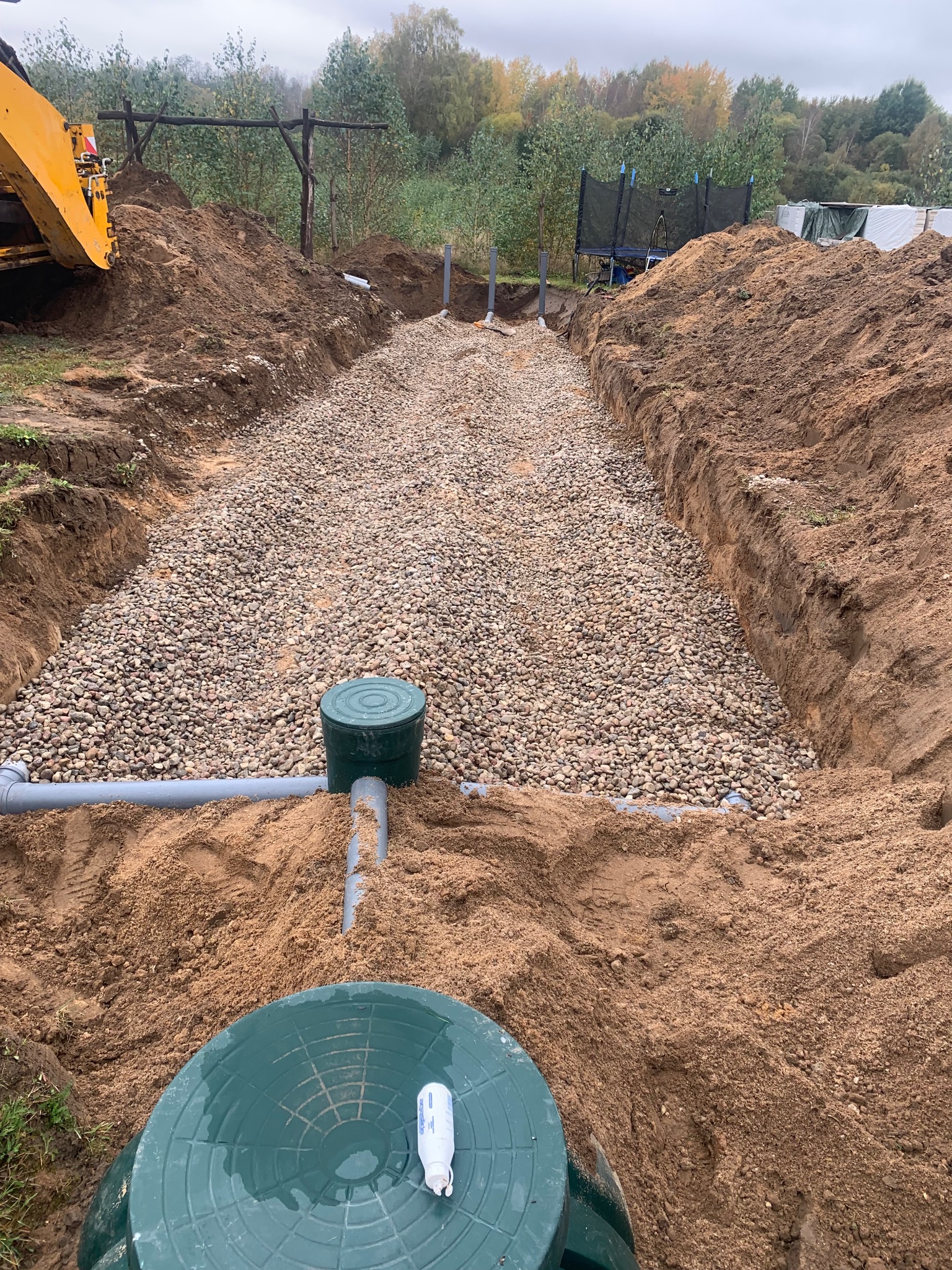 Instalacja drenażu rozsączającego z rurami i studzienkami, wypełniona żwirem w wykopie. W tle widoczny fragment koparki i drzewa.