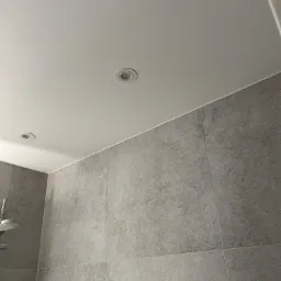 Szare płytki na ścianie łazienki z widocznymi dwoma okrągłymi lampami sufitowymi, oraz fragmentem słuchawki prysznicowej.