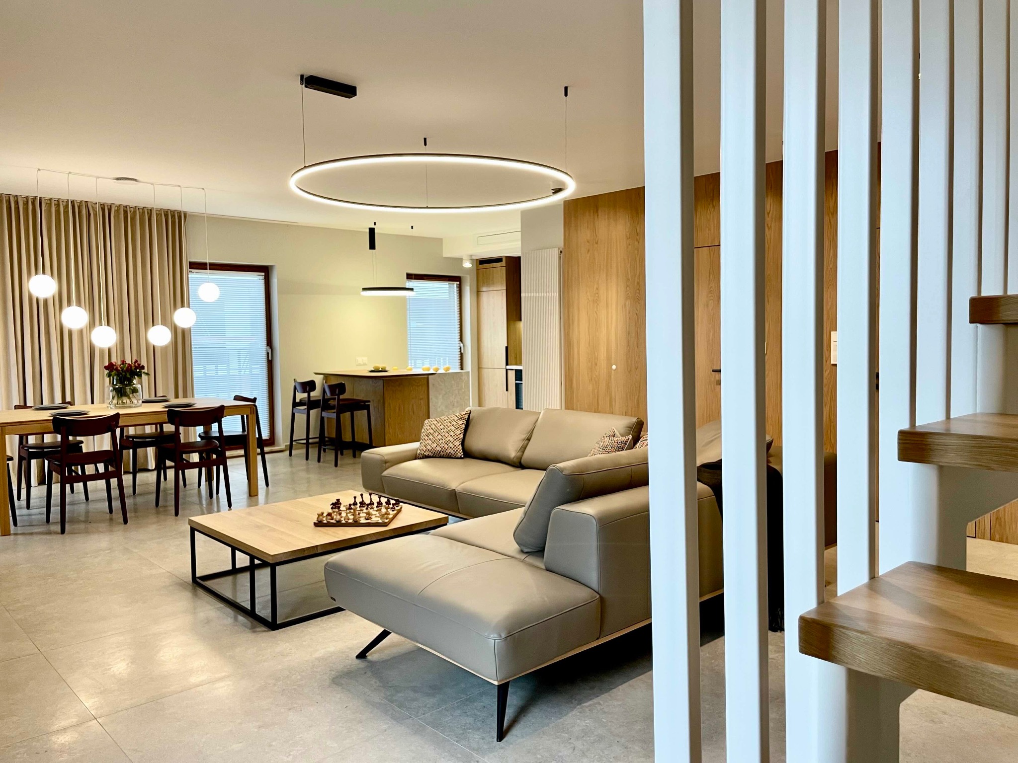 Jasne, przestronne wnętrze domu z otwartą kuchnią, jadalnią i salonem. Nowoczesne oświetlenie, drewniane akcenty i schody z białymi barierkami.