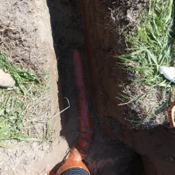 Pomarańczowa rura kanalizacyjna ułożona w wykopie ziemnym z widocznymi złączkami i karbowanym odcinkiem, obok trawa i but, zdjęcie z góry.