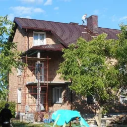 Montaż brązowego dachu z blachodachówki na dwupiętrowym domu z cegły w Koszalinie, widoczny pracownik na dachu przy kominie i rusztowanie przy ścianie.