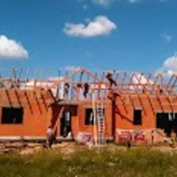 Budowa domu jednorodzinnego w trakcie montażu drewnianej konstrukcji dachu. Widoczni dwaj pracownicy na wysokości, drabina oparta o ścianę, materiały budowlane na ziemi. Niebo błękitne z białymi...