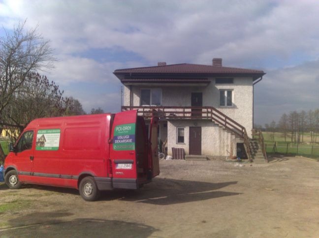 Czerwony samochód dostawczy firmy dekarskiej z otwartymi drzwiami na tle dwupiętrowego domu z brązowym dachem i drewnianymi schodami zewnętrznymi.