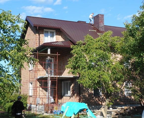 Montaż brązowego dachu z blachodachówki na dwupiętrowym domu z cegły w Koszalinie, widoczny pracownik na dachu przy kominie i rusztowanie przy ścianie.