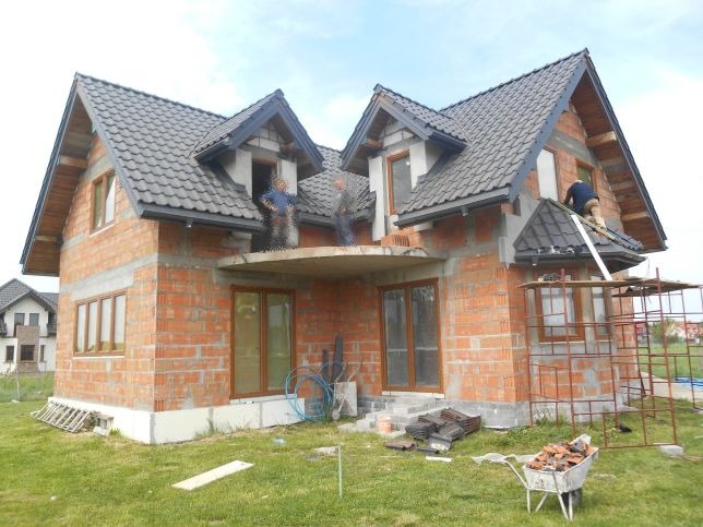 Budowa domu jednorodzinnego w trakcie, widoczni dekarze pracujący na dachu, surowy stan elewacji z cegły, rynny zamontowane, wózek z dachówkami na trawniku.