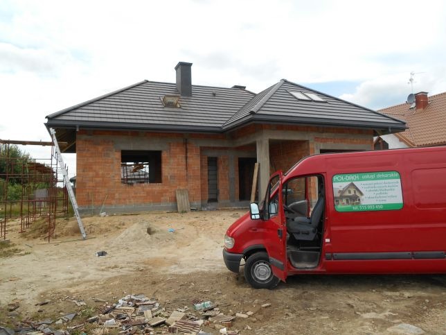 Budynek w trakcie budowy z nowym, szarym pokryciem dachowym, widoczny czerwony samochód dostawczy firmy dekarskiej z otwartymi drzwiami.