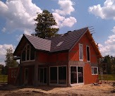 Dom w trakcie budowy, z widoczną konstrukcją dachu pokrytą ciemną dachówką, czerwone ściany w stanie surowym otwartym, białe ramy okienne, błękitne niebo z obłokami w tle.