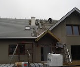 Montaż szarej dachówki na nowym domu jednorodzinnym, widoczna część dachu z kominem i oknem dachowym, dekarz pracuje na górze, rusztowanie przy ścianie budynku.