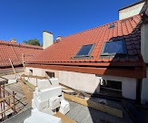 Czerwony dach z dachówki ceramicznej z dwoma oknami dachowymi, widok z rusztowania, częściowo ocieplony budynek w tle, błękitne niebo.