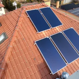 Panele słoneczne zamontowane na czerwonym dachu z dachówki ceramicznej, widok z góry na instalację i otoczenie domów jednorodzinnych.