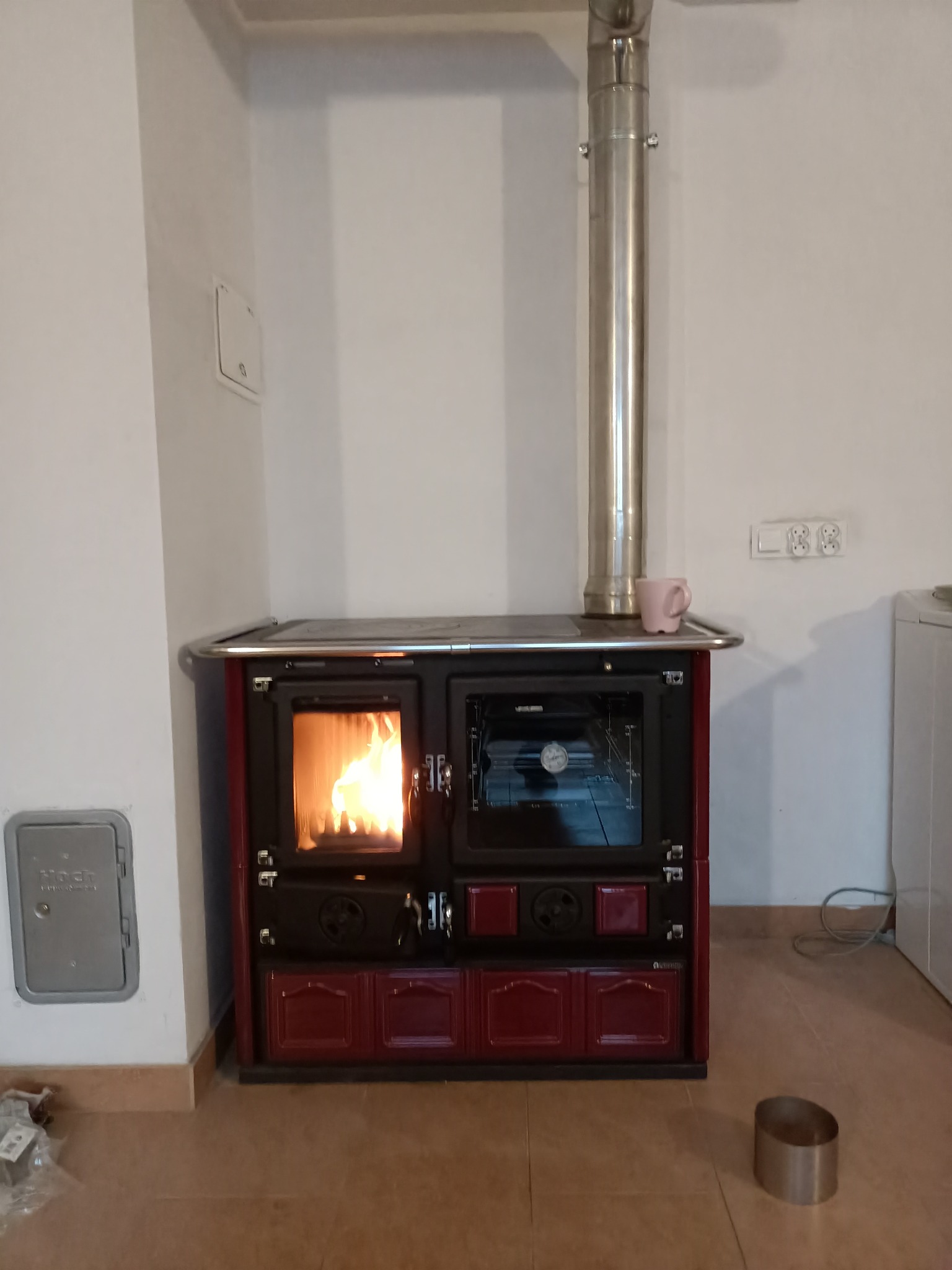 Kuchenka wolnostojąca z paleniskiem i piekarnikiem, z widocznym płomieniem, podłączona do komina ze stali nierdzewnej. Na górze kuchenki stoi różowy kubek.