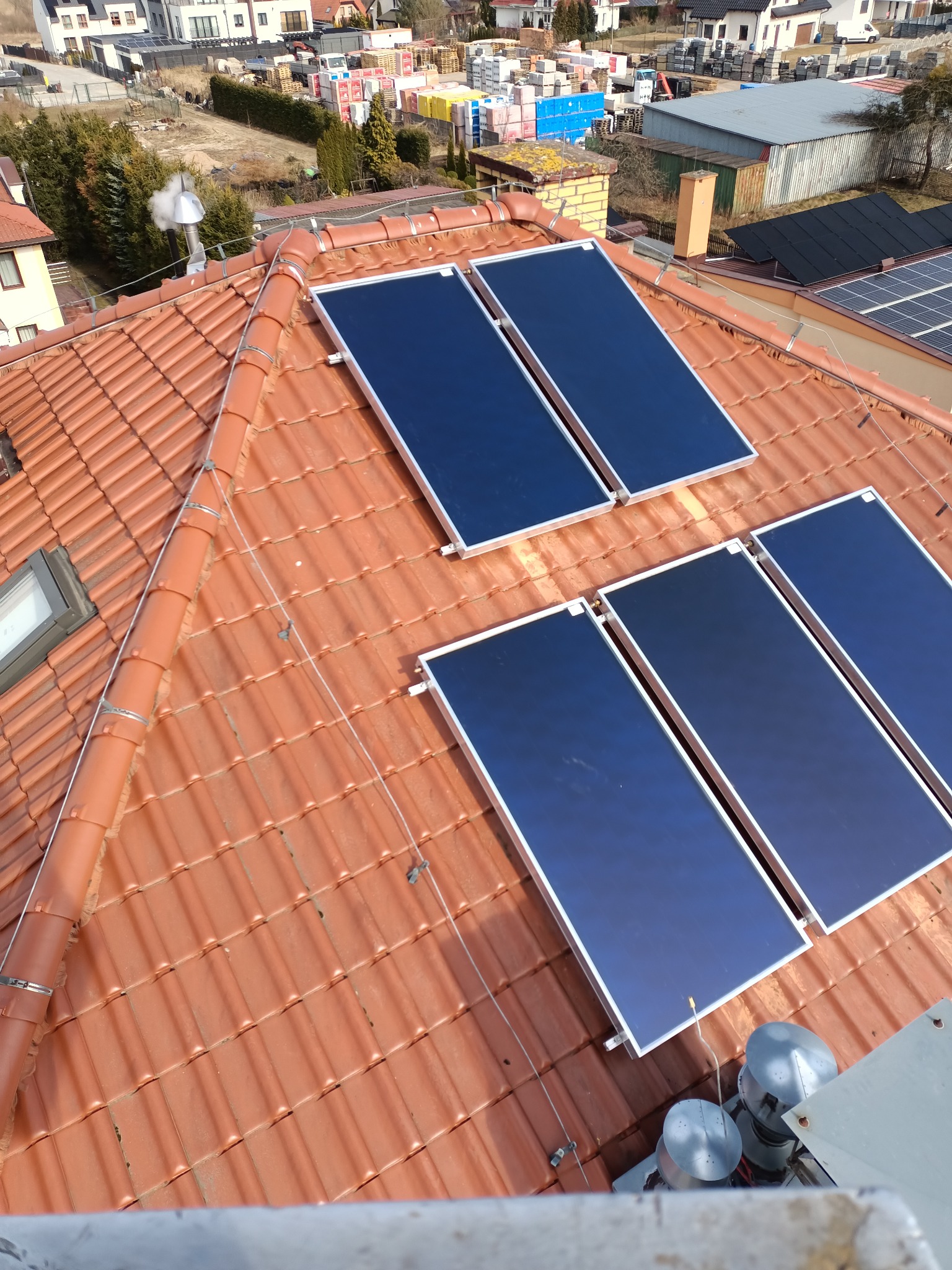 Panele słoneczne zamontowane na czerwonym dachu z dachówki ceramicznej, widok z góry na instalację i otoczenie domów jednorodzinnych.