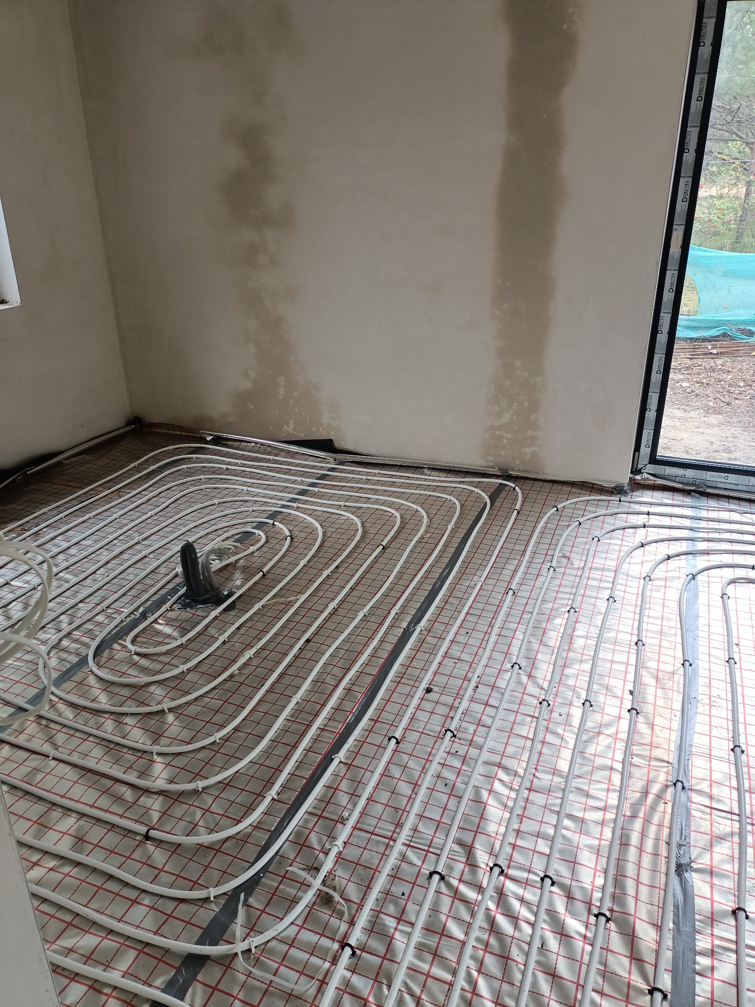 Rozłożone rury ogrzewania podłogowego na folii z siatką w surowym pomieszczeniu z oknem i wilgotnymi plamami na ścianie.