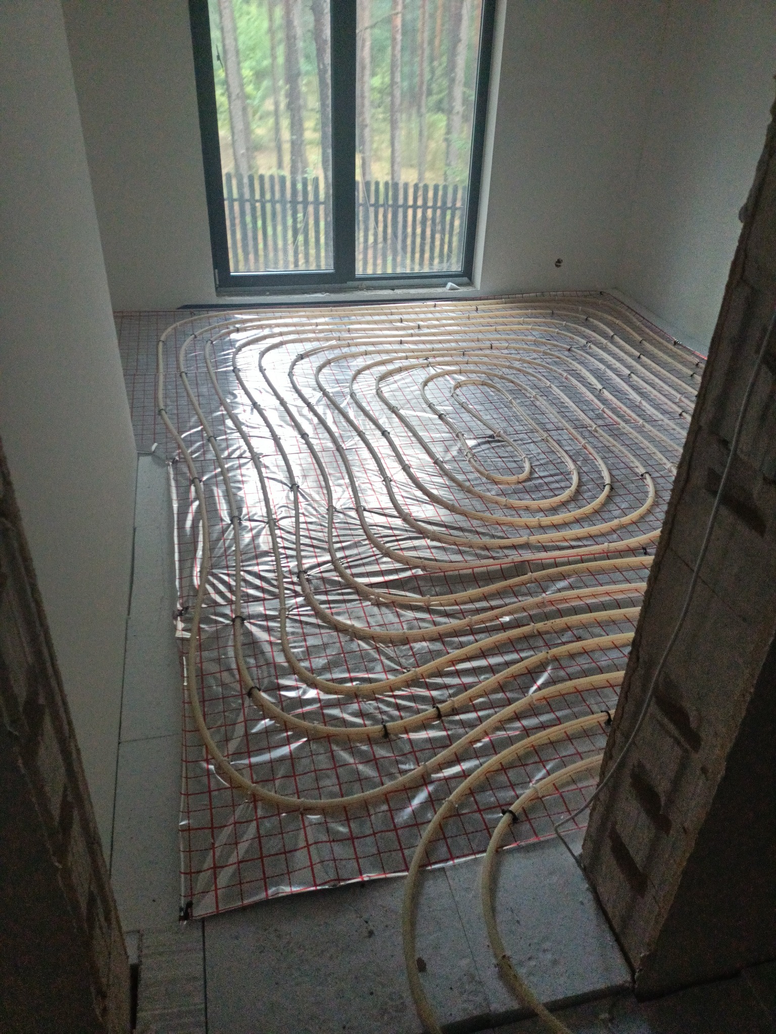 Instalacja ogrzewania podłogowego z rur w kształcie meandrów na folii z czerwoną siatką w surowym pomieszczeniu z oknem z widokiem na las.