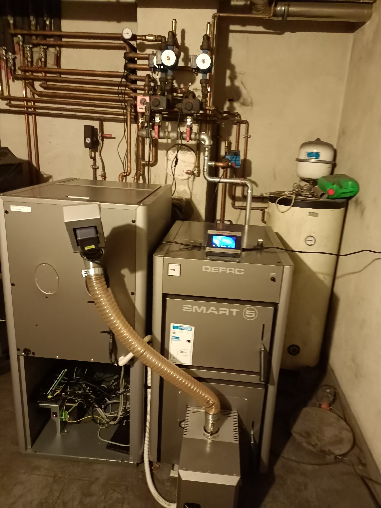 Dwa nowoczesne kotły grzewcze w kotłowni, z widocznymi rurami miedzianymi, zbiornikiem wyrównawczym i elektroniką sterującą.