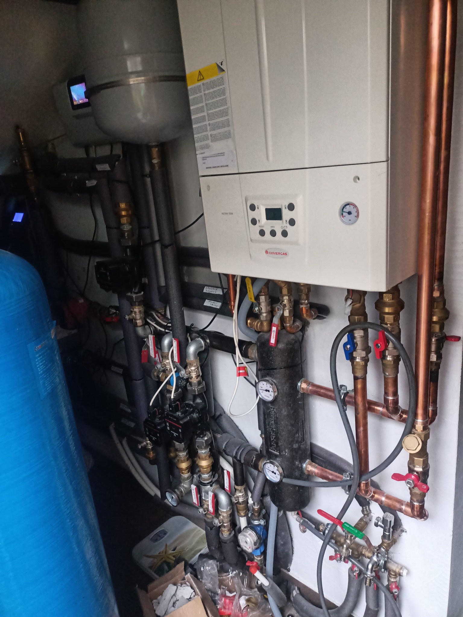 Kotłownia: biały piec gazowy Immergas z miedzianymi rurami, manometrami i zaworami, zbiornik wyrównawczy oraz niebieski zmiękczacz wody w ciasnym pomieszczeniu.