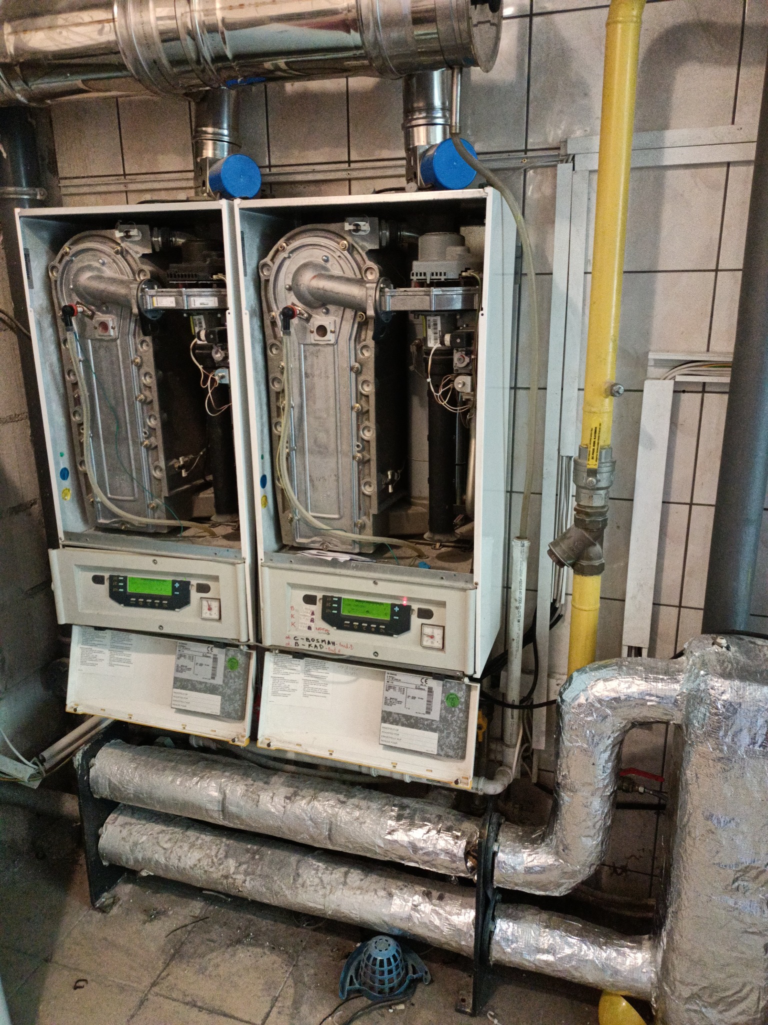 Dwa wiszące, identyczne piece gazowe z otwartymi obudowami, widoczne elementy wewnętrzne, pod nimi izolowane rury, żółta rura gazowa z zaworem po prawej stronie, tło stanowią kafelki.