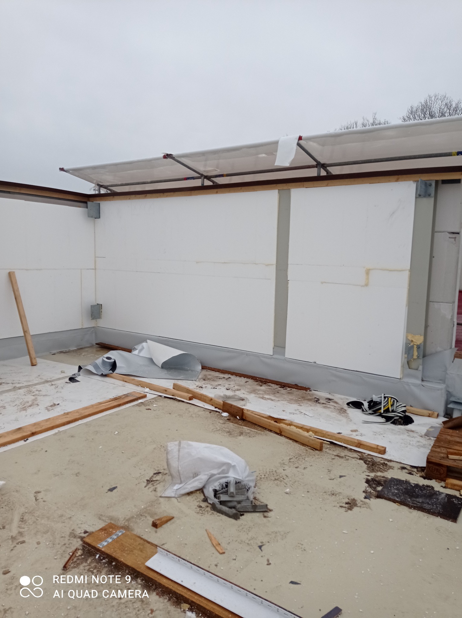 Montaż izolacji termicznej ścian budynku z widocznymi panelami styropianowymi, folią hydroizolacyjną u dołu i elementami konstrukcji dachu w trakcie budowy.