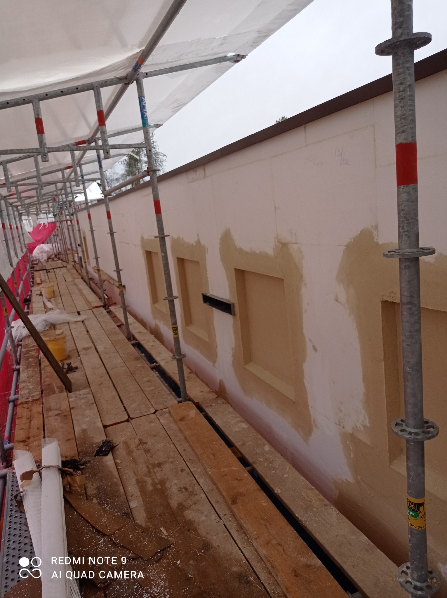 Elewacja budynku w trakcie renowacji, widoczne rusztowanie, zabezpieczenia, oraz fragmenty ściany z uzupełnionymi ubytkami wokół okien, przygotowane do dalszych prac.