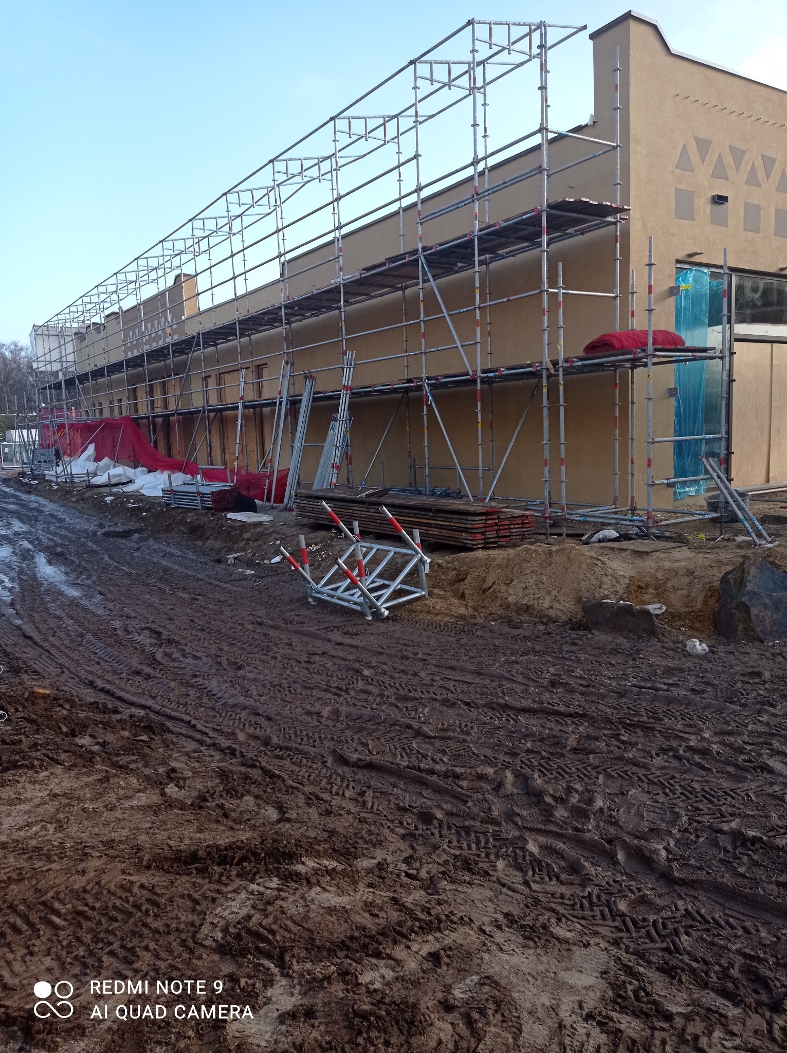 Budynek w trakcie prac elewacyjnych, widoczne rusztowania, zabezpieczenia okien folią i błotniste podłoże.