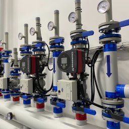 Unipam Sp. z o.o. - Rząd starannie zainstalowanych, nowoczesnych pomp obiegowych Grundfos X, z manometrami i zaworami EFAT, zamontowanych na białych rurach z niebieskimi oznaczeniami kierunku przepływu, na tle białej...