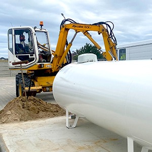 Żółta koparka Mecalac 8CX przygotowuje wykop pod biały, poziomy zbiornik na gaz na zewnątrz budynku, widoczne świeże ślady ziemi.