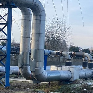 Zewnętrzna instalacja rur grzewczych, zabezpieczonych izolacją, biegnąca nad ziemią, z widocznymi elementami mocującymi i fragmentem niebieskiej konstrukcji wsporczej w tle.