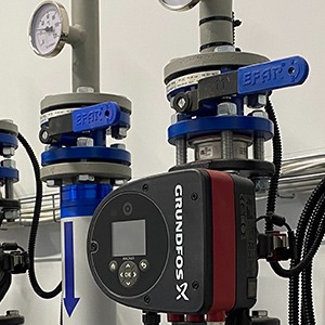 Szczegółowe ujęcie pompy obiegowej Grundfos z czerwonym akcentem, zamontowanej na instalacji z rurami i zaworami EFAR z niebieskimi uchwytami, widoczny manometr i strzałka wskazująca kierunek...