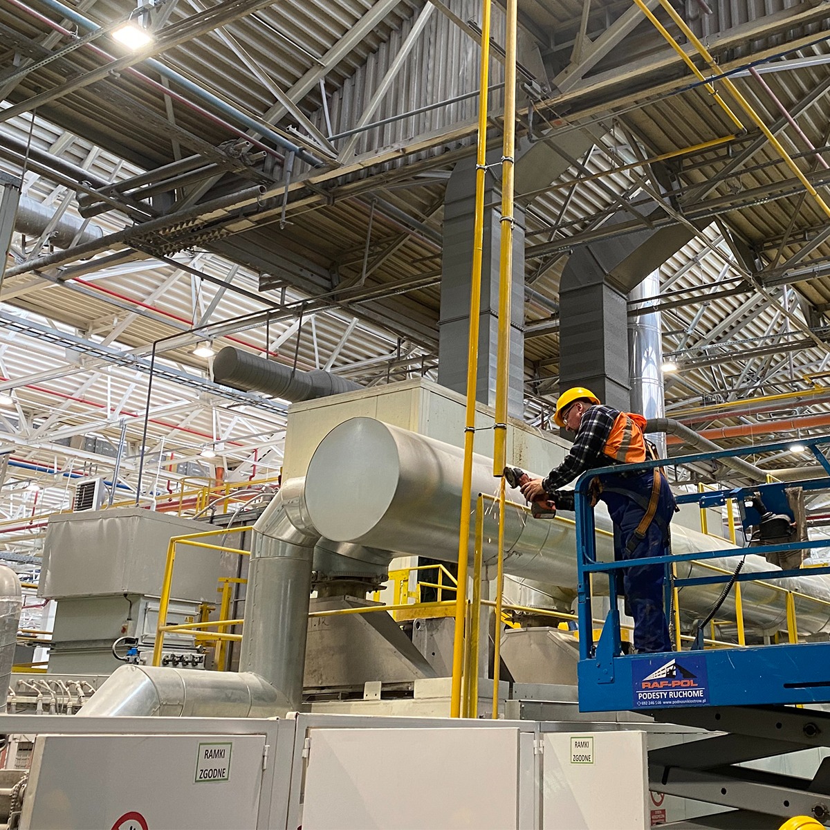 Pracownik w kasku i kamizelce ochronnej, na podnośniku koszowym, szlifuje metalową rurę instalacji przemysłowej w hali fabrycznej z widoczną konstrukcją dachu.