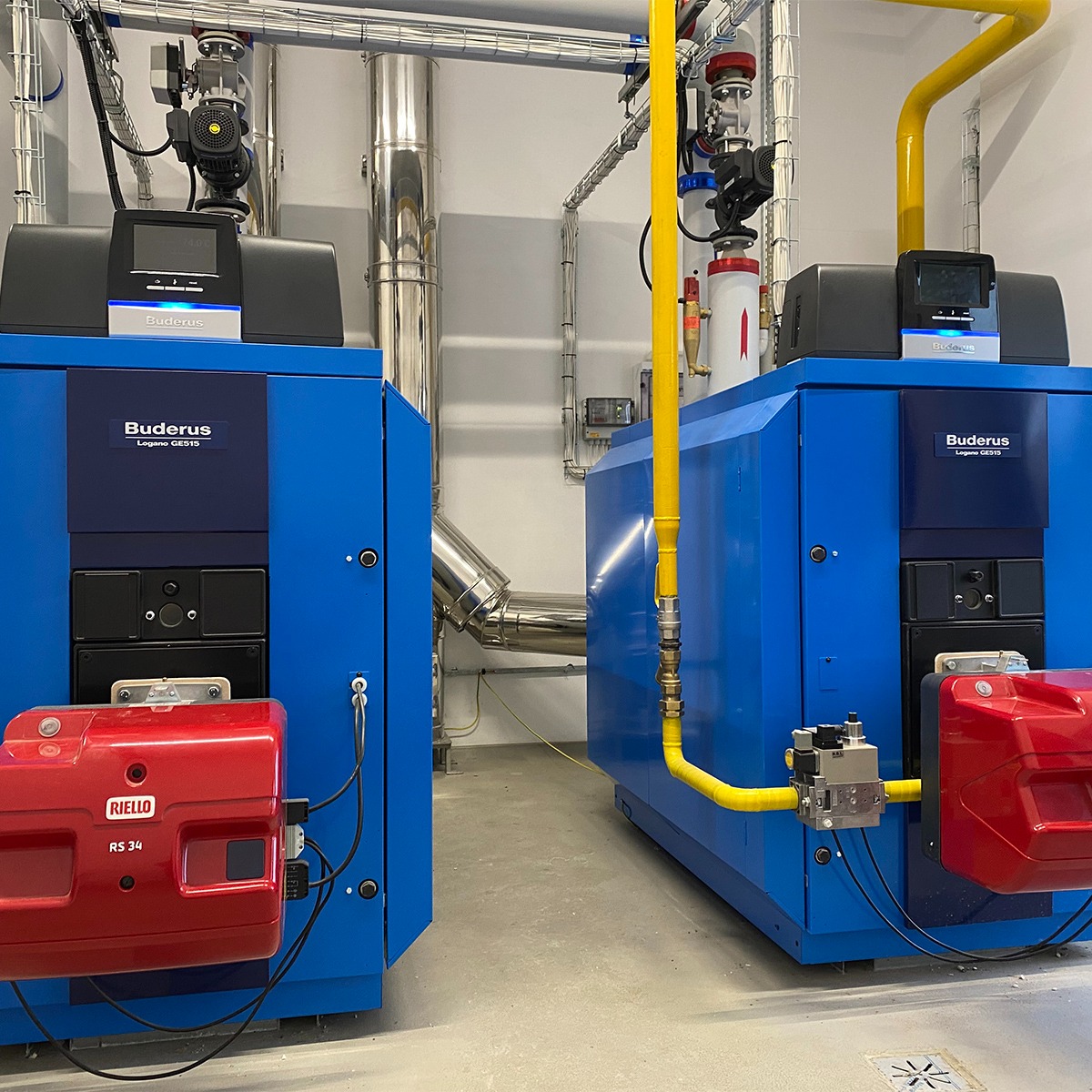 Dwa niebieskie, przemysłowe kotły gazowe marki Buderus Logano GE515 z czerwonymi palnikami Riello RS 34, połączone żółtymi rurami gazowymi w sterylnej, białej kotłowni z widocznymi elementami...