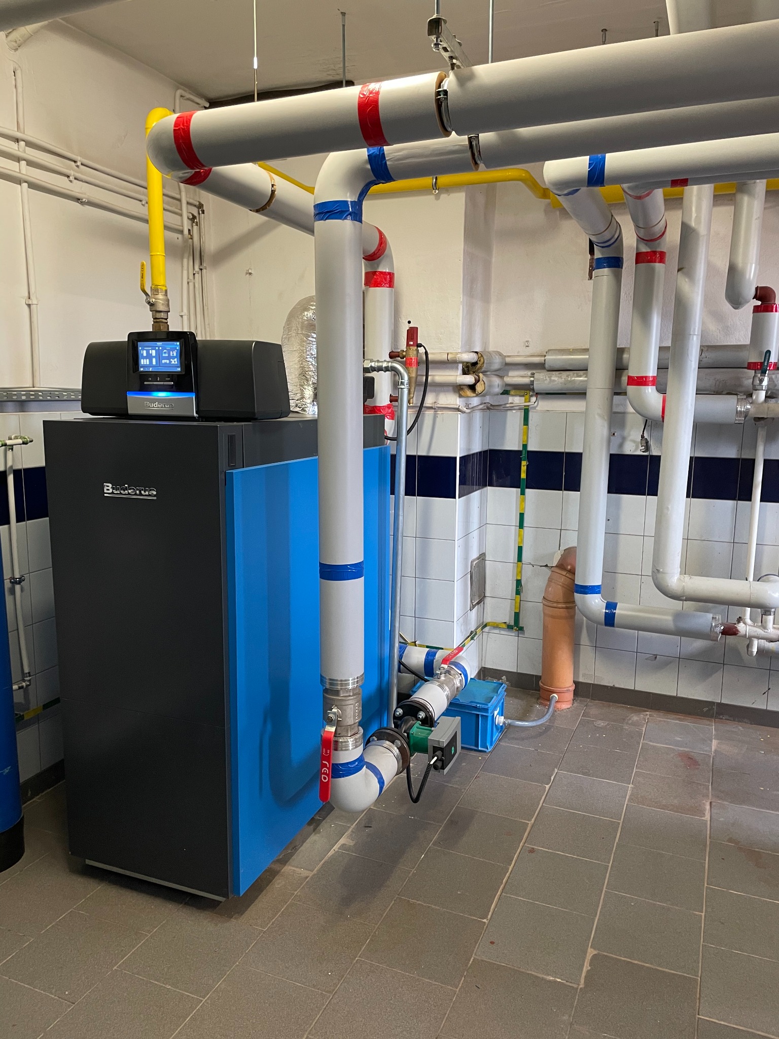 Nowoczesny, niebiesko-czarny piec gazowy Buderus z dotykowym panelem sterowania w kotłowni z rurami oznaczonymi kolorowymi taśmami.