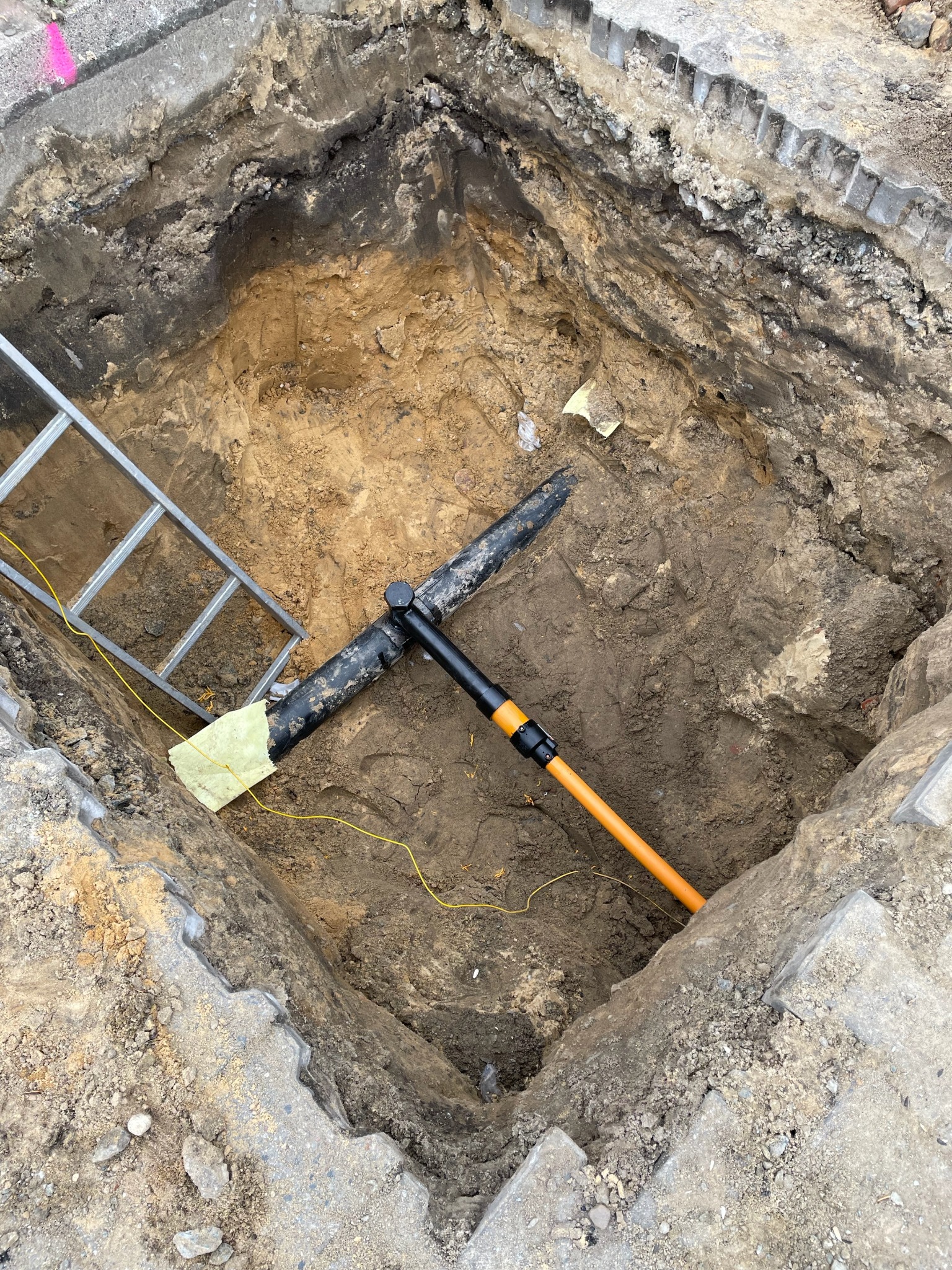 Wykop w ziemi z widoczną rurą gazową owiniętą izolacją, metalową drabinką i narzędziem z pomarańczową rękojeścią.