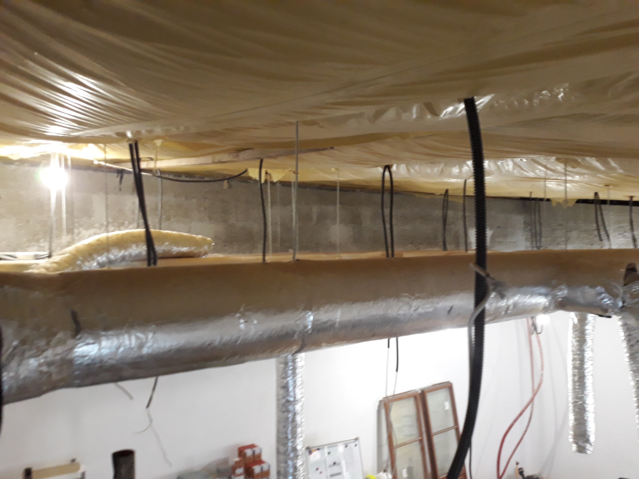 Instalacja izolacji termicznej i przewodów wentylacyjnych pod stropem, widoczne elementy mocujące i folia ochronna na suficie.