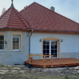 Nowoczesny dom z czerwonym dachem z dachówki karpiówki i drewnianym tarasem, elewacja w trakcie wykończenia.