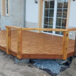 Świeżo wybudowany, drewniany taras z balustradą przy budynku z elewacją z jasnych płytek, widoczna sterta kamieni budowlanych w tle.