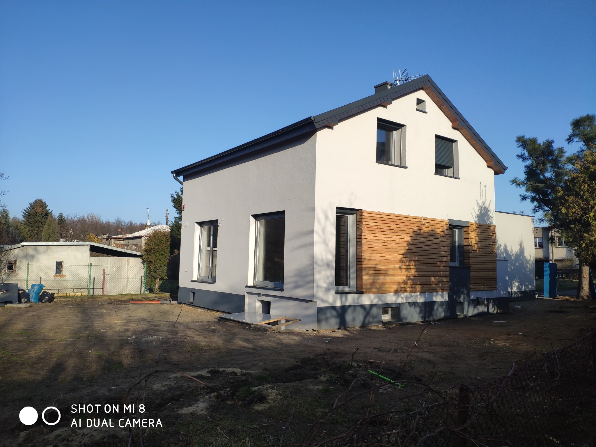 Nowoczesny dom jednorodzinny z szarą elewacją, fragmentem drewnianej okładziny i ciemnoszarym dachem, sfotografowany w słoneczny dzień na tle błękitnego nieba.