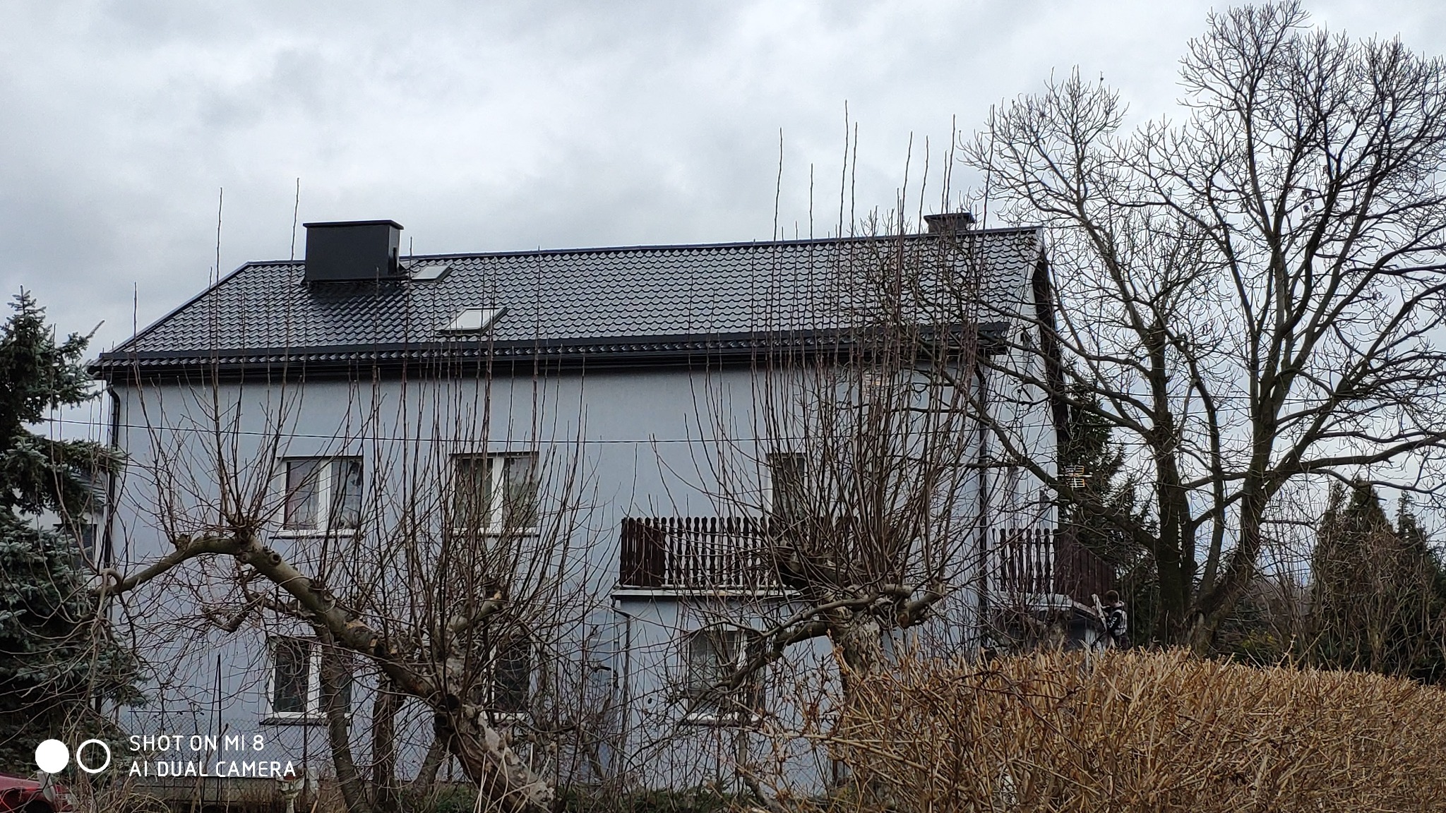 Dwupiętrowy dom z szarą elewacją i nowym, ciemnoszarym dachem z dachówki, widoczny spod gałęzi drzew. Dach z kominem i oknami dachowymi, balkon z drewnianą balustradą.