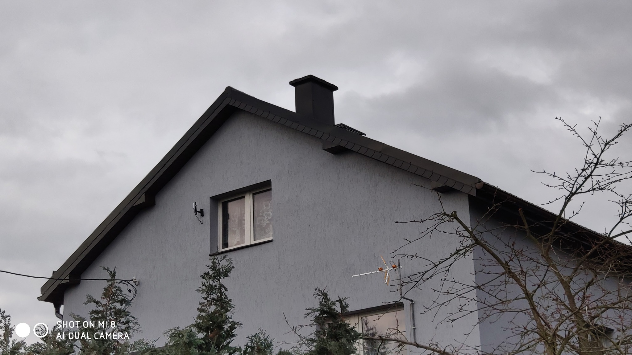 Szary dom jednorodzinny z ciemną dachówką, kominem i elewacją o fakturze baranka, widoczny z dołu na tle pochmurnego nieba.