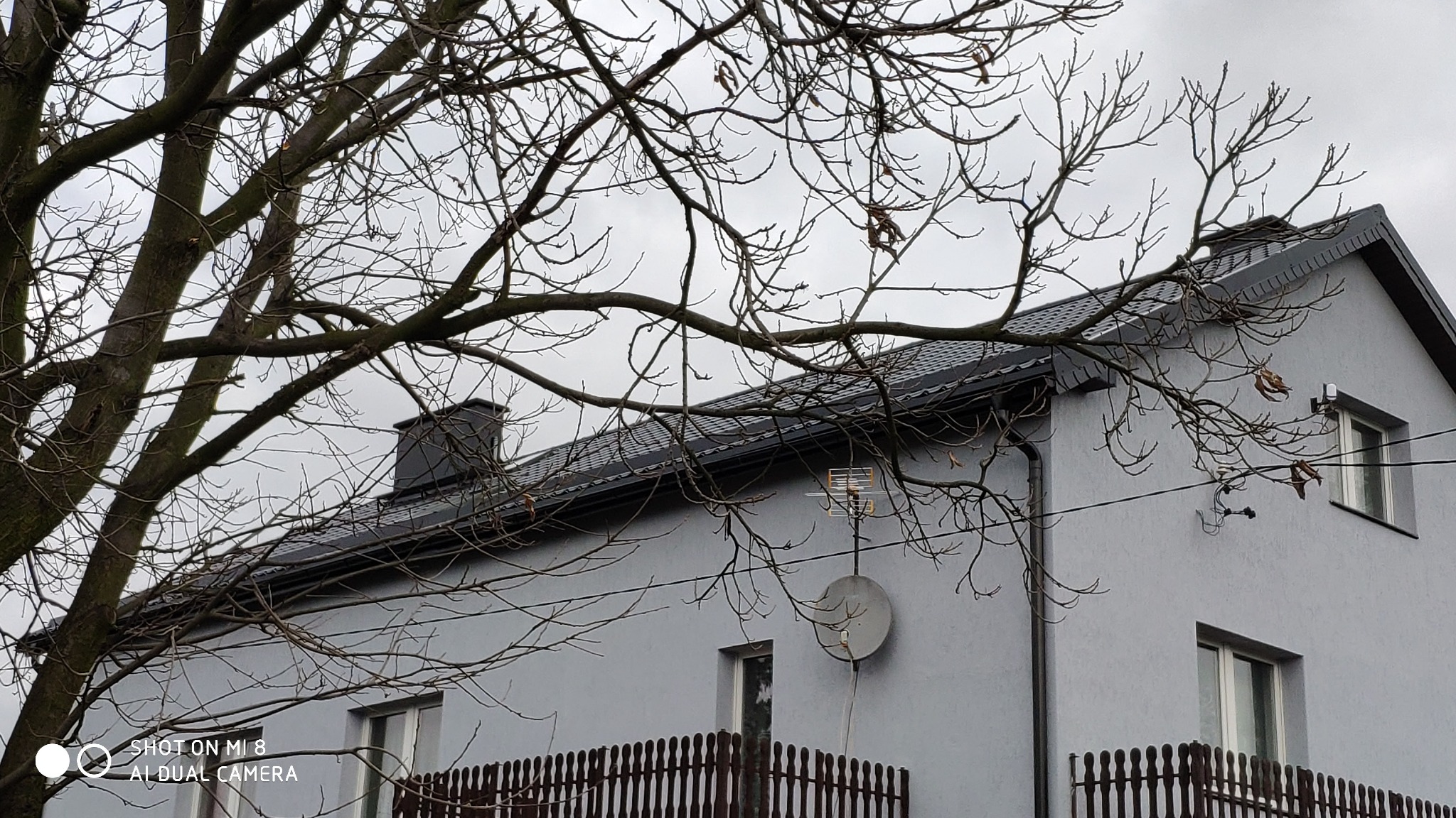 Dom z nowym, ciemnoszarym dachem z blachodachówki, widoczny komin, antena satelitarna i drzewo bez liści na pierwszym planie, pochmurne niebo.